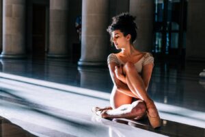 Młoda baletnica w cielistym kostiumie i w pointach siedzi na podłodze z ugiętymi nogami w kolanach, pada na nią snop światła słonecznego - jakby z okna.