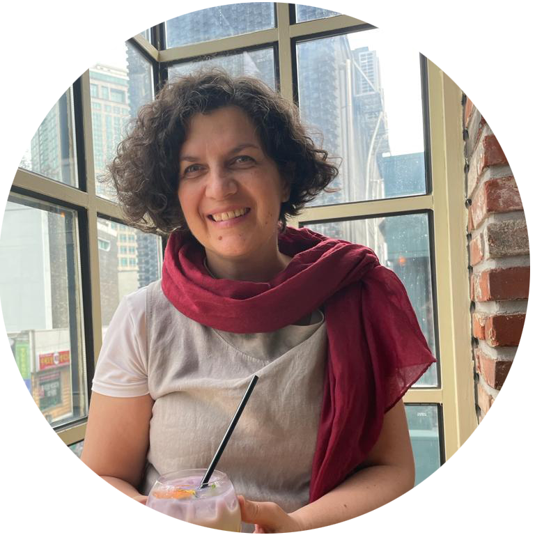 Zdjęcie portretowe uśmiechniętej kobiety w średnim wieku, ciemnobrązowe, półdługie, kręcone włosy. Ubrana w lnianą, beżową tunikę i lekki, bordowy szal wokół szyi. W rękach trzyma szklankę z mrożoną herbatą. W tle okno kawiarni.