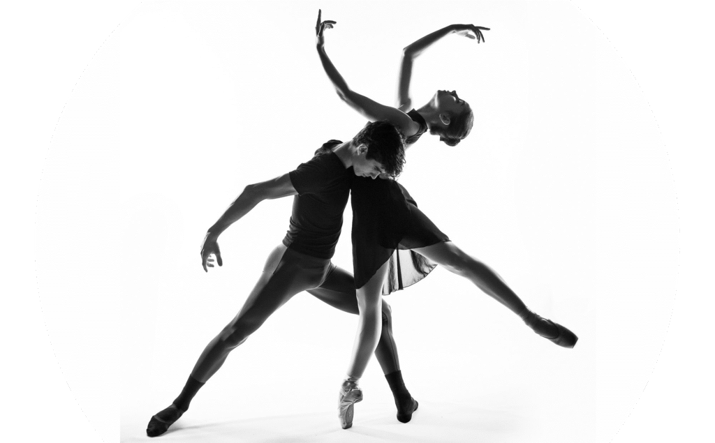 Młody chłopak i młoda dziewczyna w pozie baletowej. Tancerz trzyma tancerkę w pół patrząc w dół, tancerka na pointach, jedna noga wyciągnięta do tyłu, głowa wygięta do tyłu, ręce ponad głową. Zdjęcie czarno-białe.